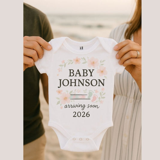 White baby onesie with 'Baby Johnson arriving soon 2026' text with floral and baby footprint graphic design
