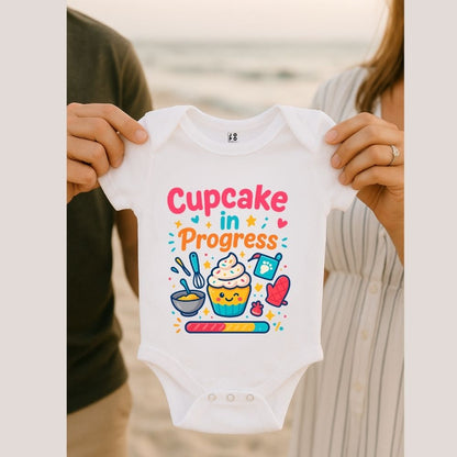 White baby onesie with 'Cupcake in Progress' text and cartoon-style cupcake design with loading bar graphic